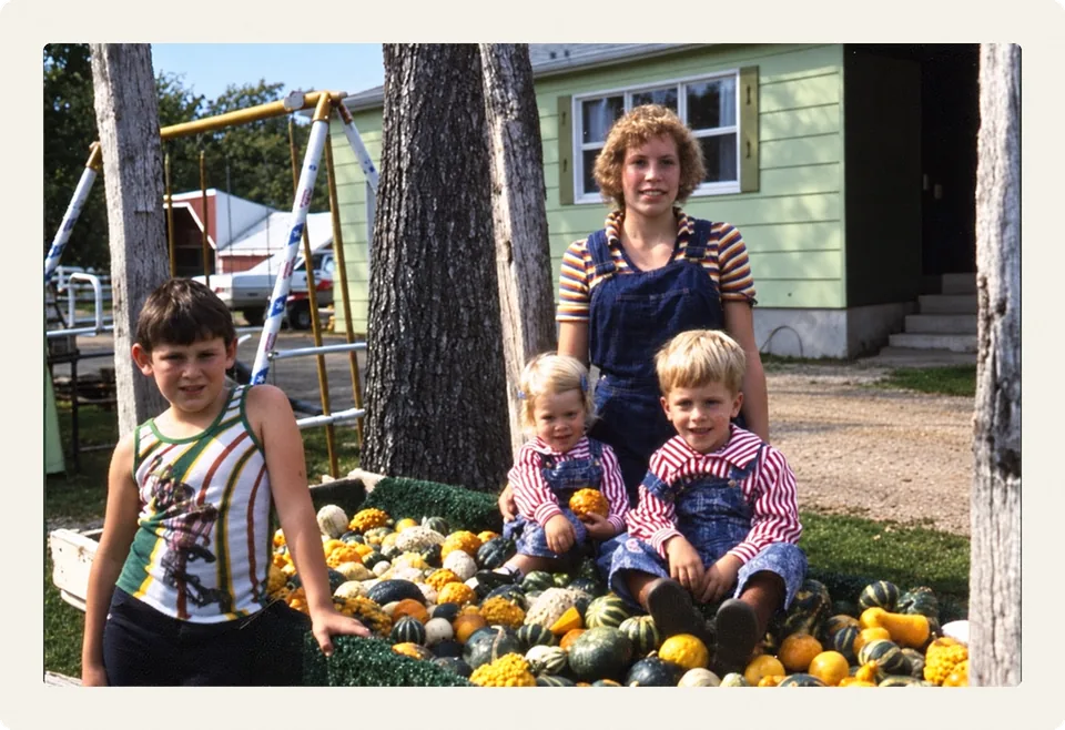 Cindy in overalls with Todd, Troy and Tania on a wagon of gourds by the green farmhouse