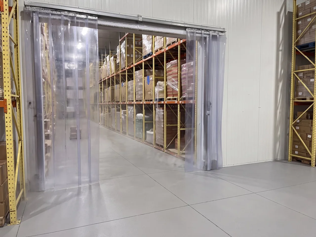 Clear plastic strip curtains across the doorway into a freezer room, with loaded yellow and orange pallet racks and a little cold haze beyond